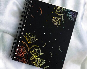 Könnte beinhalten: Ein schwarzes Spiralnotizbuch mit einem Regenbogen-Blumendesign. Die Blumen sind in einem Regenbogen von Farben umrandet und über den schwarzen Einband verteilt.