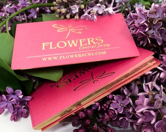 Puede incluir: Pila de tarjetas de visita rosadas con letras en relieve de oro y bordes dorados. El texto de la tarjeta dice "FLOWERS Central Jersey WWW.FLOWERSCNJ.COM".