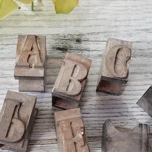May include: A set of six vintage metal letterpress printing blocks, each featuring a different letter of the alphabet: A, B, C, D, E, and I. The blocks are arranged on a white wooden surface.