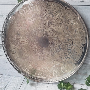 May include: A large, round silver-toned metal serving tray featuring an intricate, embossed scrollwork pattern across its surface and a decorative, perforated border along the outer edge. The tray has a polished, reflective finish throughout.