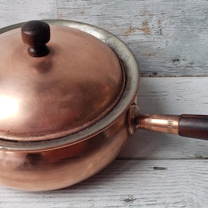 May include: A round copper saucepan with a matching lid, featuring a dark brown wooden handle and a small dark brown wooden knob on the lid. The metal surface has a polished finish, and the rim of the pan shows a silver-toned metallic band.