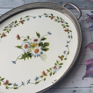 May include: An oval serving tray with a silver-toned metal rim featuring a pierced gallery edge and two handles. The centre is a white surface decorated with a central bouquet of daisies and a surrounding wreath of small blue and orange flowers.