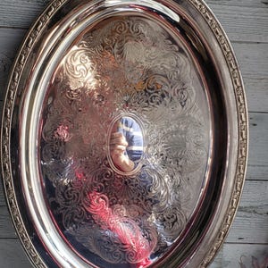 May include: An oval silver-toned serving tray featuring ornate embossed scrollwork and floral patterns across the surface. The tray includes two decorative handles at the top and bottom and a raised border with a repeating geometric design.