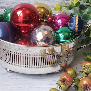 May include: A silver bowl with a decorative design and feet, filled with colourful Christmas baubles. The bowl has two handles and is filled with baubles in red, green, blue, gold, and silver.