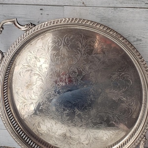 May include: A round silver-toned serving tray featuring an intricate engraved floral pattern across the centre surface. The tray has a raised, rope-style border and a decorative pierced gallery edge, complemented by two ornate, curved metal handles.