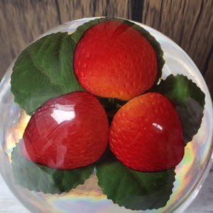 May include: A clear glass orb containing three red strawberries on green leaves. The strawberries have a glossy finish and are arranged in a circular pattern. The background is a wooden surface.