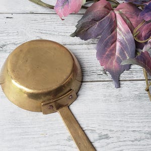 May include: A vintage brass saucepan with a long handle and a small, round bowl. The handle has a hole for hanging. The saucepan is set against a white wooden background with purple and pink leaves.
