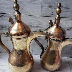 May include: Two antique brass and copper coffee pots with ornate designs. The pots have a rounded body with a spout and a handle on each side. The tops of the pots have a lid with a finial.