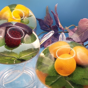 May include: Three clear glass teacups with fruit and leaf designs encased in resin. One cup features a yellow fruit, another a red berry, and the third, oranges. The cups sit on a light blue surface.