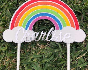 May include: A rainbow cake topper with a white cloud base and the name "Charlise" written in silver glitter. The rainbow is made of red, orange, yellow, green, blue, indigo, and violet paper with glitter accents.