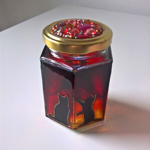 May include: A hexagonal glass jar with a gold lid decorated with red and iridescent rhinestones. The jar is filled with a red liquid and features a black silhouette of two cats on the front.