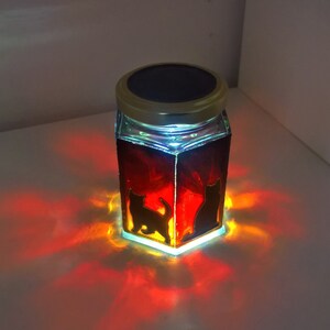 May include: A glass jar with a solar powered lid, featuring a silhouette of two cats. The jar is filled with a red liquid and glows orange when lit.