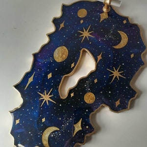 May include: A resin tray with a dark blue and purple galaxy design. The tray is decorated with gold glitter, stars, moons, and a gold rim. The tray has two gold handles with clear glass inserts.