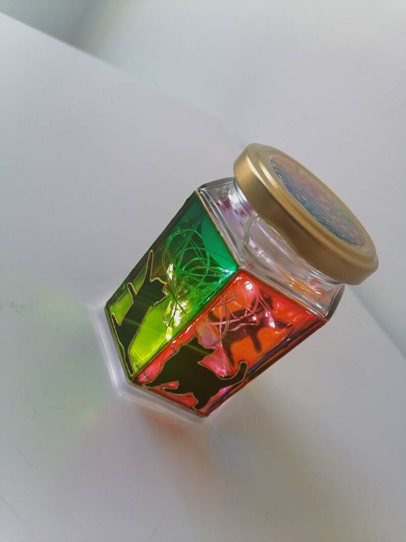 May include: A glass jar with a gold lid, decorated with a rainbow-colored design and black silhouettes of animals. The jar is filled with fairy lights.