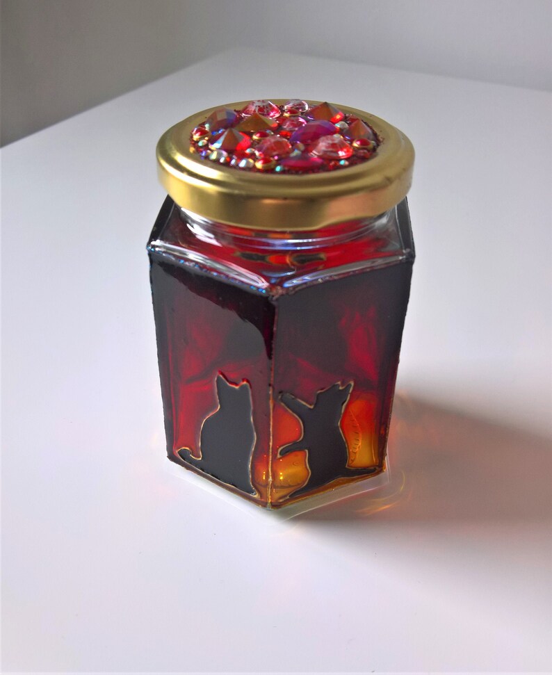 May include: A hexagonal glass jar with a gold lid decorated with red and iridescent rhinestones. The jar is filled with a red liquid and features a black silhouette of two cats on the front.