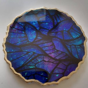 Puede incluir: Un posavasos redondo de resina azul y morado con un borde dorado y un diseño negro similar al de una vidriera.