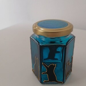 May include: A small, hexagonal glass jar with a gold lid. The jar is filled with a teal blue liquid and has black cat silhouettes painted on the sides.