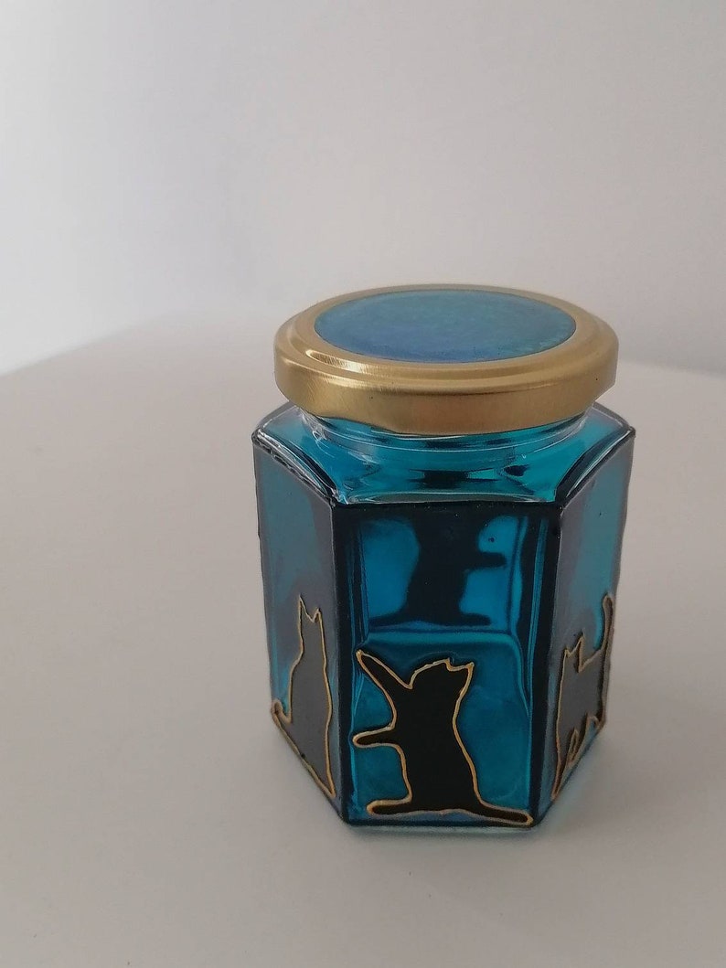 May include: A small, hexagonal glass jar with a gold lid. The jar is filled with a teal blue liquid and has black cat silhouettes painted on the sides.