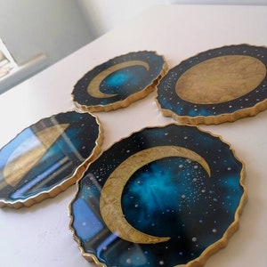 Puede incluir: Conjunto de cuatro posavasos redondos de resina con borde dorado y diseño de galaxia. Cada posavasos presenta una fase lunar diferente en oro.