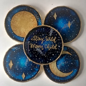 May include: Set of four round coasters with a gold rim. Each coaster features a different celestial design with gold accents on a dark blue background. One coaster has a full moon, another has a crescent moon, and two have a starry night sky with diamond-shaped stars. The center coaster has the text "Stay Wild Moon Child" in gold lettering.