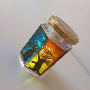May include: A glass jar with a gold lid, decorated with colorful stained glass panels featuring black cat silhouettes. The jar is filled with fairy lights, creating a warm and whimsical glow.