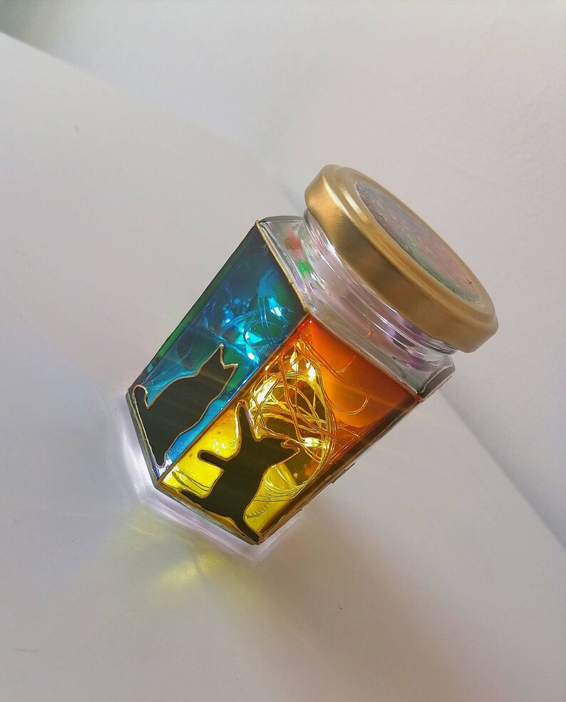 May include: A glass jar with a gold lid, decorated with colorful stained glass panels featuring black cat silhouettes. The jar is filled with fairy lights, creating a warm and whimsical glow.