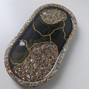 May include: An oval-shaped resin tray with a black base and gold glitter accents. The tray is decorated with gold lines and filled with iridescent glitter. The edges are lined with glitter, creating a decorative and functional piece.