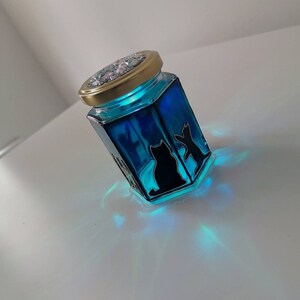 May include: A blue glass jar with a gold lid and a glittery top. The jar has a silhouette of two cats inside. The jar is lit from within, casting a blue glow.