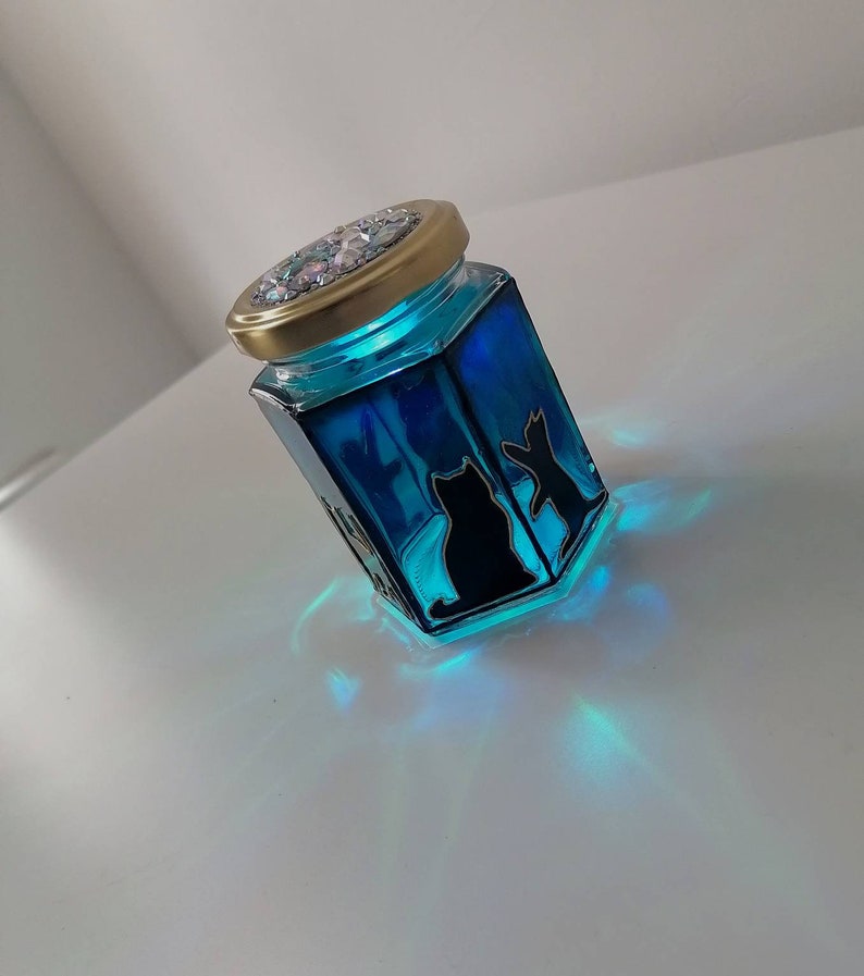 May include: A blue glass jar with a gold lid and a glittery top. The jar has a silhouette of two cats inside. The jar is lit from within, casting a blue glow.