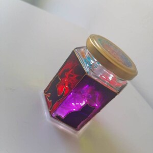 May include: A glass jar with a gold lid, decorated with black cat silhouettes and stained glass in shades of red and purple. The jar is filled with colorful glitter and lights up when lit.