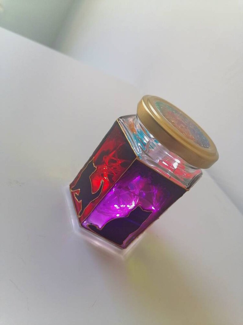 May include: A glass jar with a gold lid, decorated with black cat silhouettes and stained glass in shades of red and purple. The jar is filled with colorful glitter and lights up when lit.