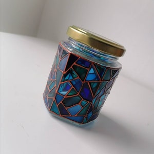 May include: A glass jar with a gold lid decorated with a mosaic of blue, green, and black geometric shapes outlined in copper.