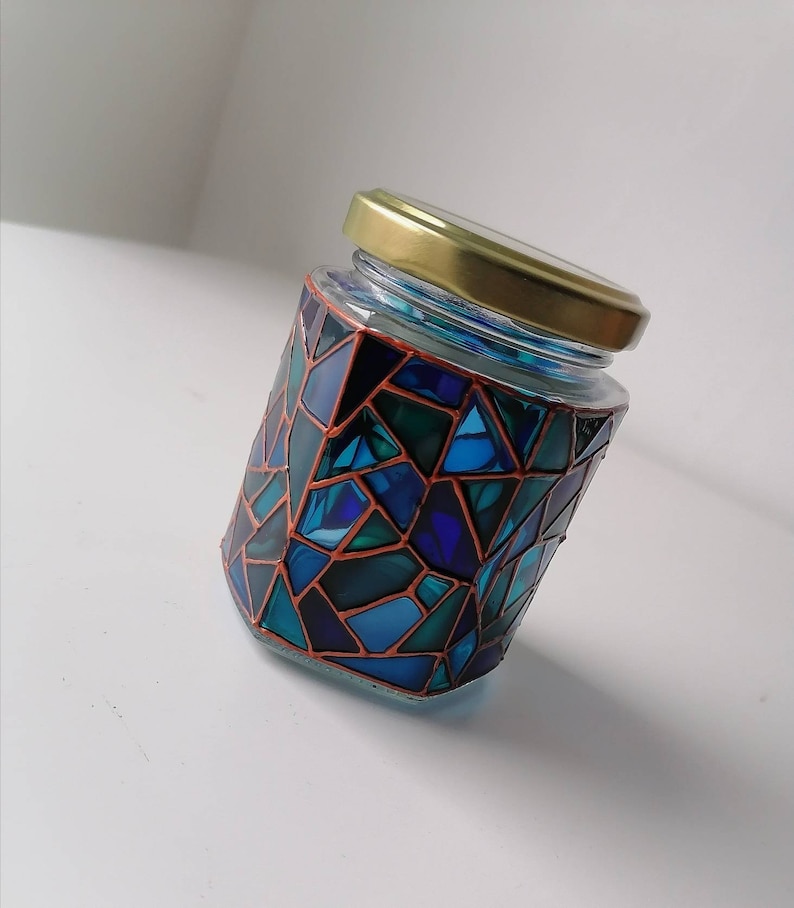 May include: A glass jar with a gold lid decorated with a mosaic of blue, green, and black geometric shapes outlined in copper.