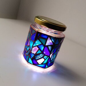 May include: A glass jar with a gold lid decorated with a mosaic of blue, purple, and teal glass pieces. The jar is lit from within, creating a colorful glow.