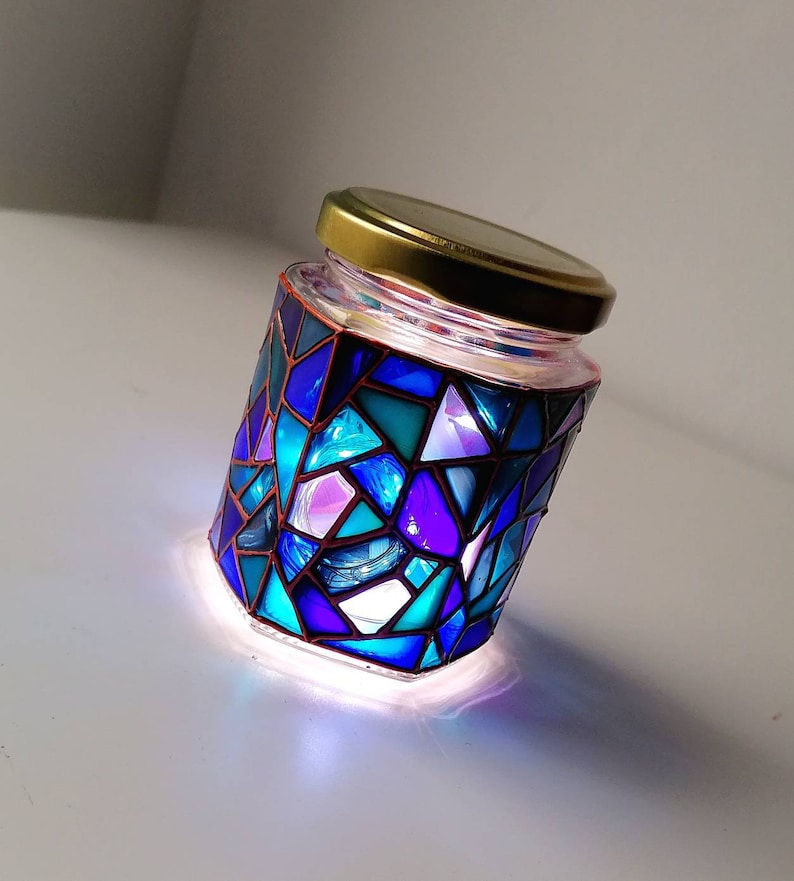 May include: A glass jar with a gold lid decorated with a mosaic of blue, purple, and teal glass pieces. The jar is lit from within, creating a colorful glow.