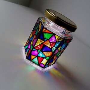 May include: A glass jar with a gold lid, decorated with colorful stained glass-like designs. The jar is illuminated from within, casting a rainbow of colors on the surface.