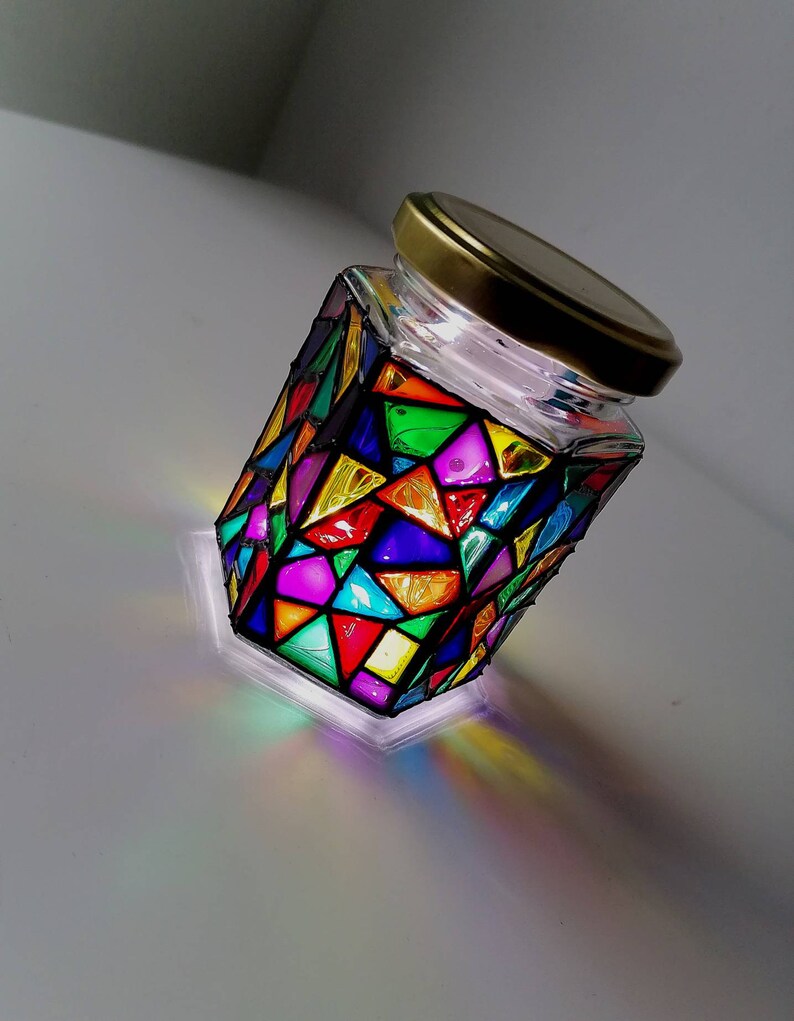 May include: A glass jar with a gold lid, decorated with colorful stained glass-like designs. The jar is illuminated from within, casting a rainbow of colors on the surface.