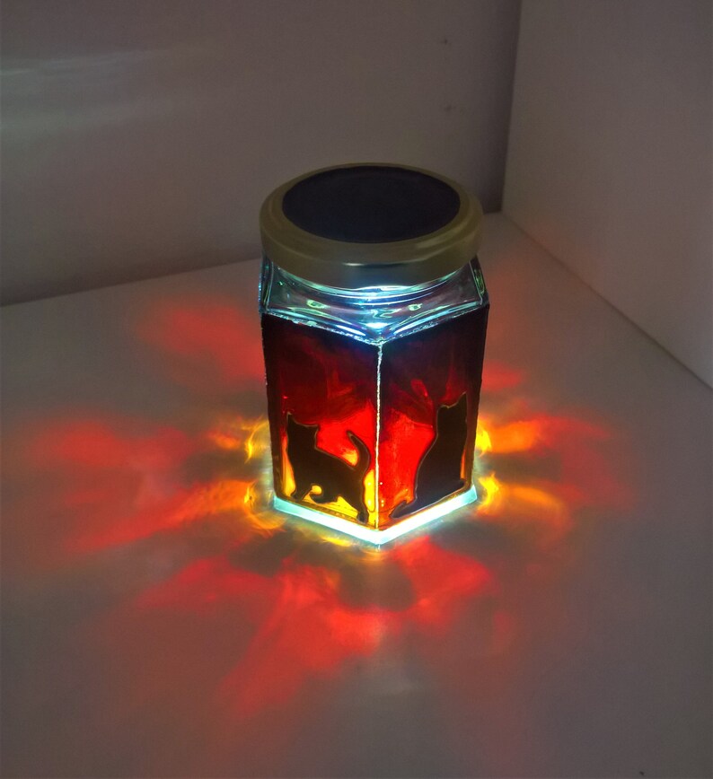 May include: A glass jar with a solar powered lid, featuring a silhouette of two cats. The jar is filled with a red liquid and glows orange when lit.