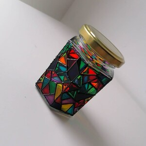 May include: A glass jar with a gold lid decorated with colorful geometric shapes and black lines. The jar is filled with a clear liquid.