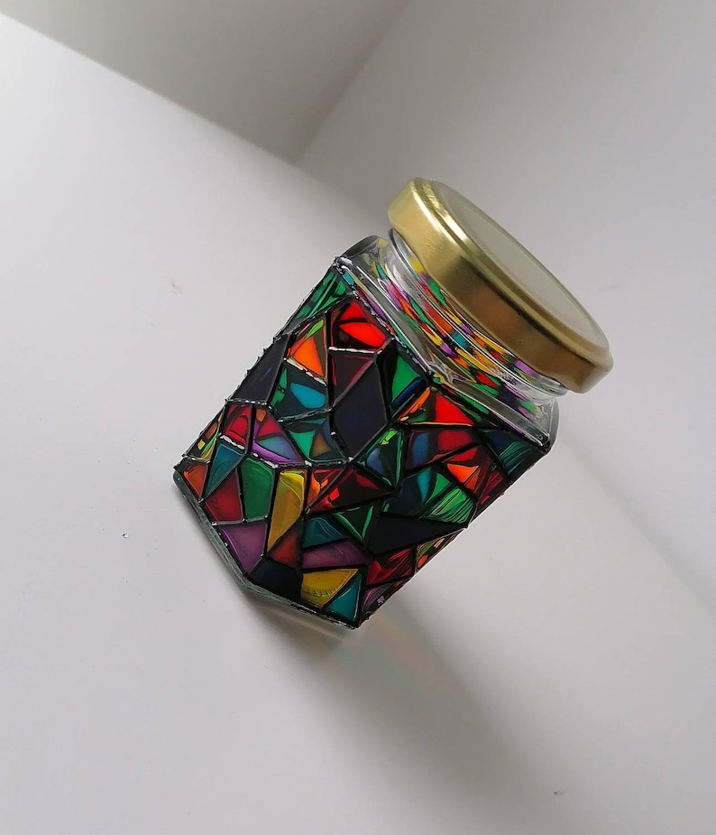 May include: A glass jar with a gold lid decorated with colorful geometric shapes and black lines. The jar is filled with a clear liquid.