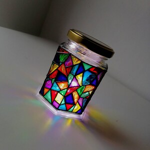 May include: A glass jar with a gold lid, decorated with colorful stained glass-like geometric patterns. The jar is lit from within, casting a rainbow glow.