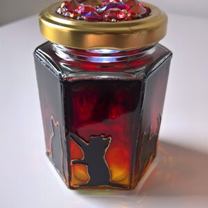 May include: A hexagonal glass jar with a gold lid and a red and orange ombre effect. The lid is decorated with iridescent rhinestones. The jar features black silhouette images of cats.