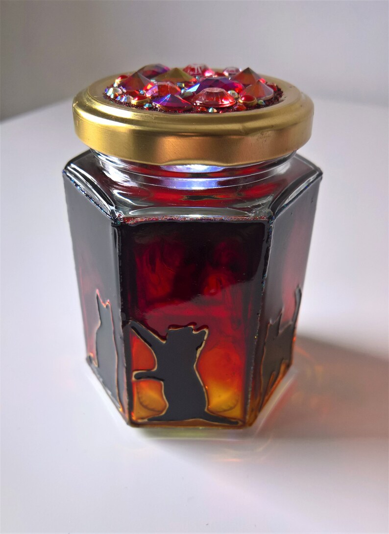 May include: A hexagonal glass jar with a gold lid and a red and orange ombre effect. The lid is decorated with iridescent rhinestones. The jar features black silhouette images of cats.
