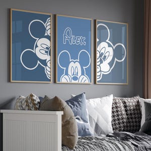 Könnte beinhalten: Drei gerahmte blaue Wandkunstwerke mit Mickey Mouse. Ein Stück trägt den Namen "Alex" in weißer Schrift. Die Kunst hängt über einem weißen Tagesbett mit Kissen und einer gemusterten Decke. Zwei Weidenkörbe stehen vor dem Bett.