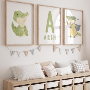 Op de afbeelding: Drie ingelijste kunstprints met een kinderkamerthema. De prints tonen een groene alligator, een grote groene letter "A" met de naam "Aiden" en een superheld alligator. Onder de prints staat een houten opbergbank met witte bakken en kussens.