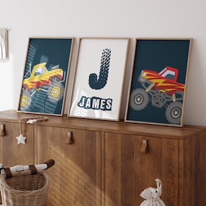 May include: Three framed prints with a blue background and monster truck illustrations. The center print has the name "JAMES" in white letters. The left print shows a yellow monster truck and the right print shows a red monster truck.