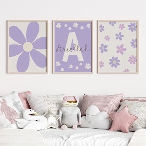 May include: Three framed prints with a purple flower design. The center print features the letter "A" and the name "Ariella" in cursive. The prints are hanging on a white wall above a white bed with pillows.