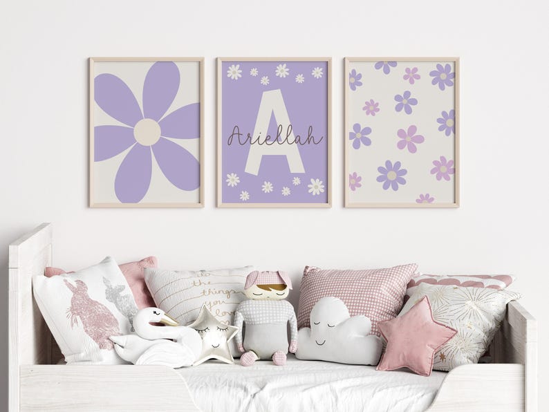 May include: Three framed prints with a purple flower design. The center print features the letter "A" and the name "Ariella" in cursive. The prints are hanging on a white wall above a white bed with pillows.