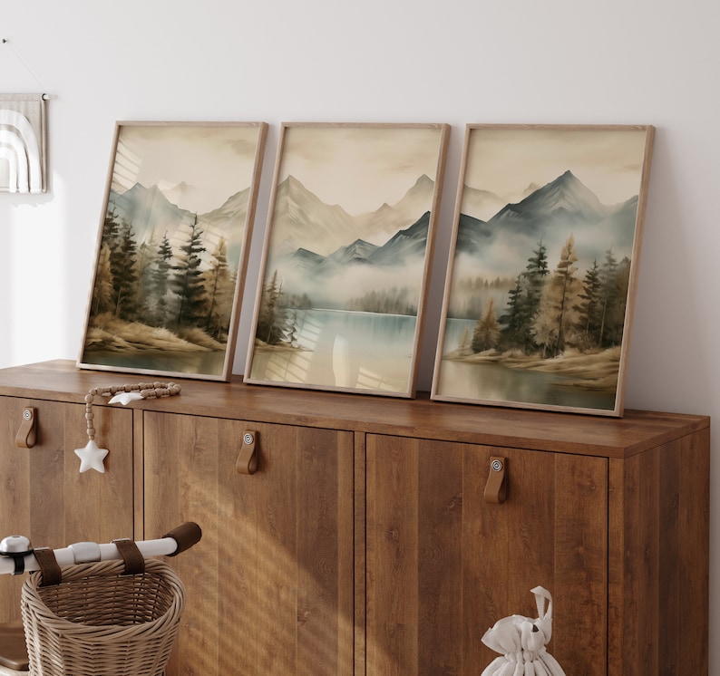 May include: Three framed watercolor paintings depicting a misty mountain landscape with a lake in the foreground. The paintings are in shades of brown, blue, and green, and are hung on a wooden wall.