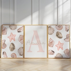 Puede incluir: Tres impresiones artísticas enmarcadas con temática de playa. Dos impresiones presentan ilustraciones de acuarela de conchas marinas y estrellas de mar en tonos beige y rosa. La impresión central muestra el nombre "Amillia" y una gran letra "A" en rosa pálido.